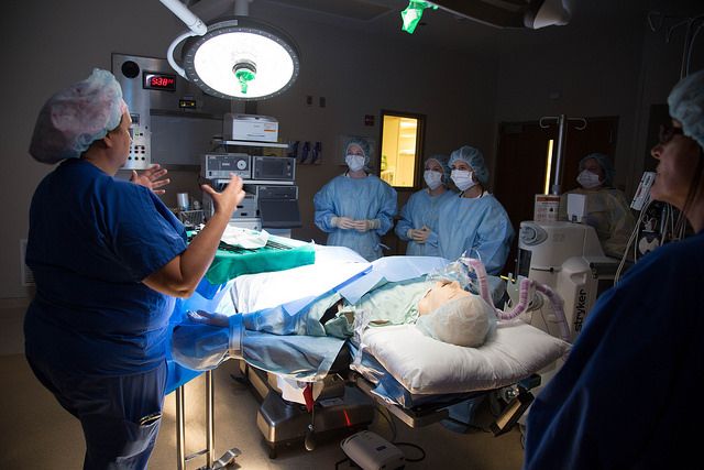 Nursing Academy students in medical scrubs listen to a surgeon as she instructs them over a medical manikin in a darkened room.